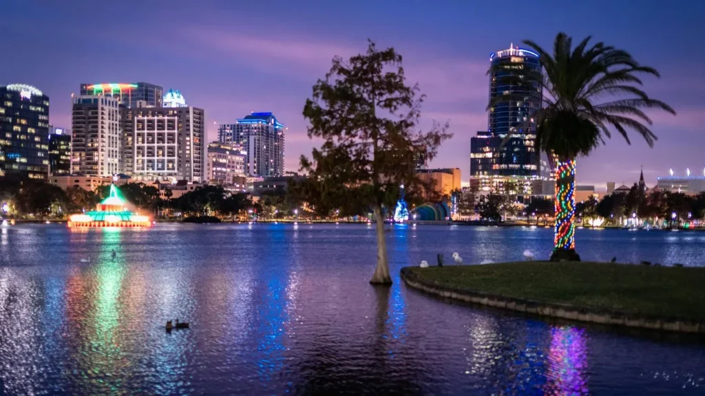 wilight view of Lake Eola Park in Orlando with a cypress tree, palm tree wrapped in holiday lights, and city skyline in the background
