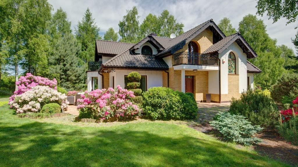 Exterior view of a two-story brick and stucco home with a dark roof, surrounded by a lush green lawn and vibrant, blooming pink rhododendrons in a beautifully landscaped garden.