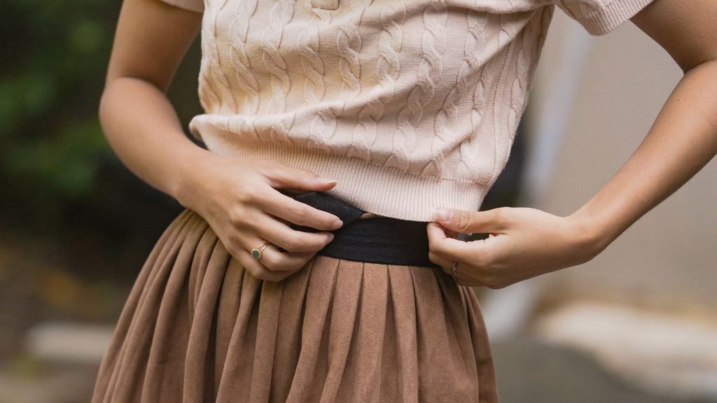 Close-up of a beige cable-knit sweater tucked into a pleated tan skirt with a black waistband