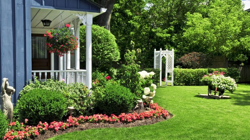 A charming blue house with a white columned front porch, adorned with hanging flower baskets, looking out over a lush green lawn, red border flowers, and a white garden trellis.