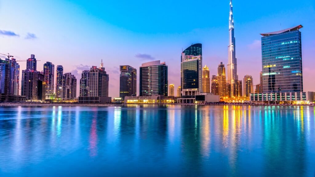 Downtown Dubai skyline lit up at dusk, with the Burj Khalifa and surrounding towers reflecting on the calm water in the foreground.