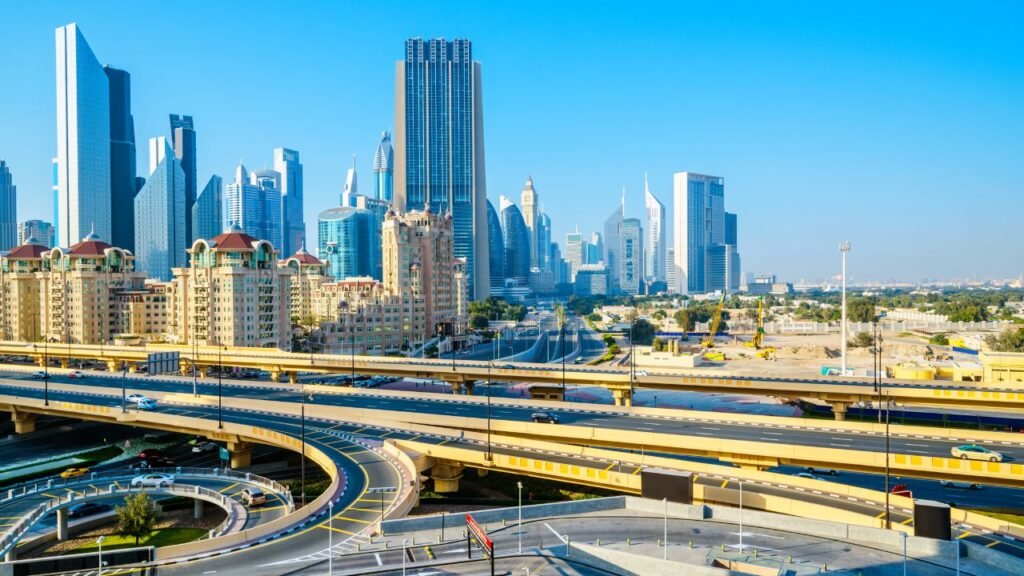 A curving multi-lane highway interchange in Dubai with the city's skyscraper skyline, including the Emirates Towers, visible under a clear blue sky