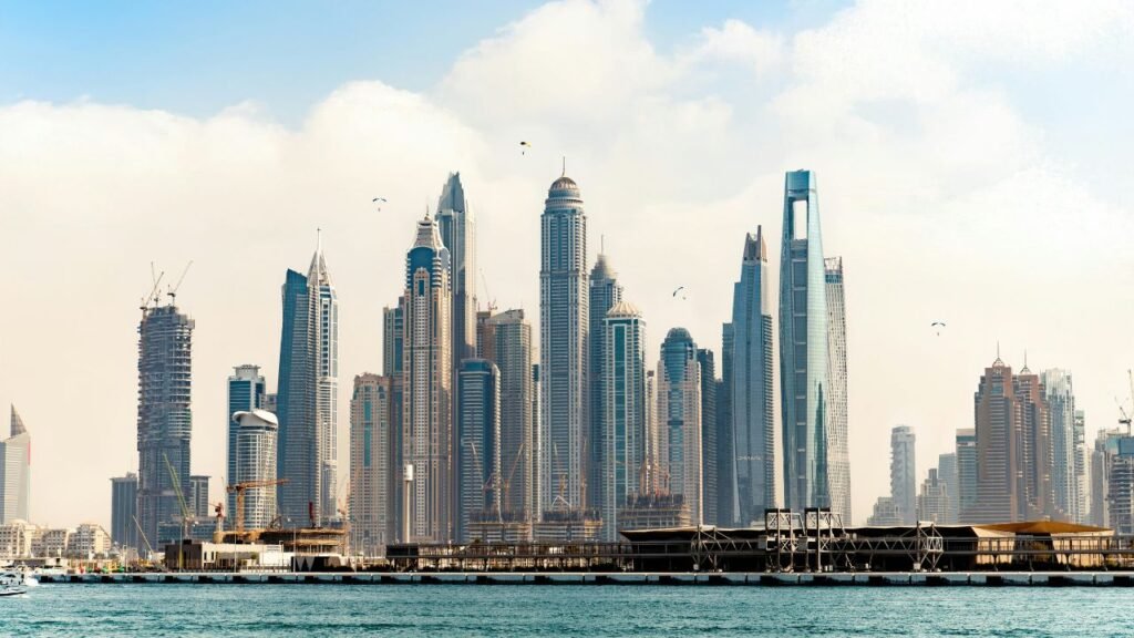 Dubai Marina skyline with a cluster of high-rise towers along the waterfront under a partly cloudy sky, with paragliders visible in the air.