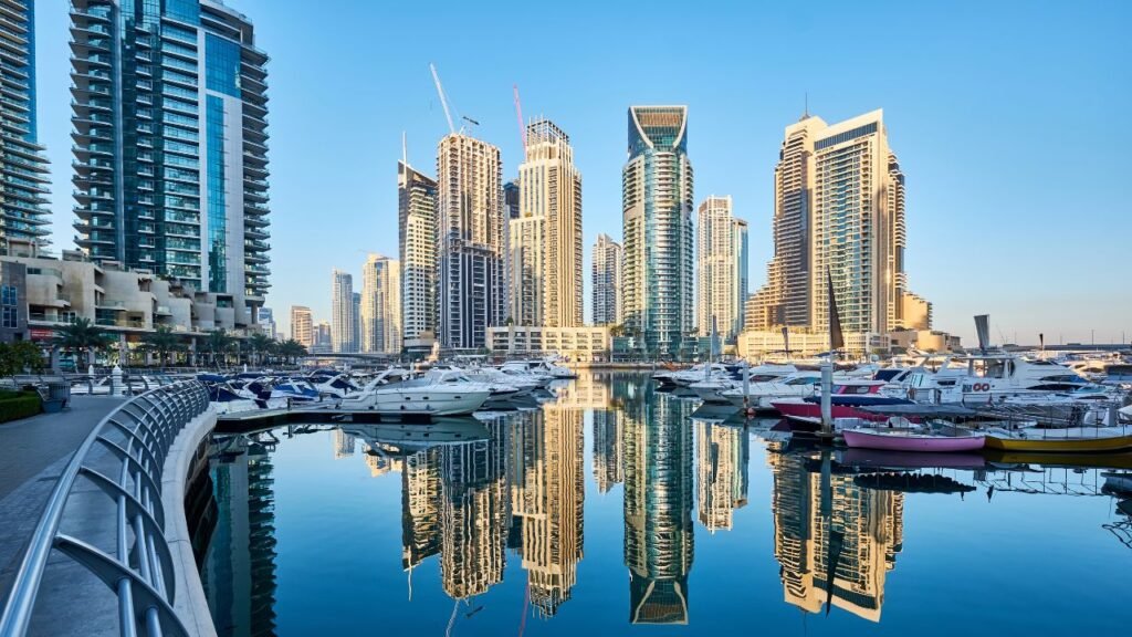 Yachts and boats docked in Dubai Marina with high-rise towers reflected in the still water during early morning light.