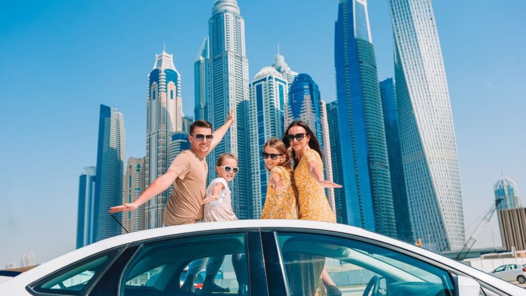 A family of four standing through a car's sunroof, posing happily with the Dubai Marina skyscrapers in the background on a sunny day.
