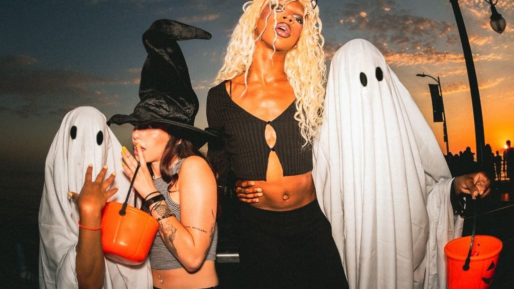 Group of friends in ghost, witch, and wig Halloween costumes trick-or-treating at the beach during sunset