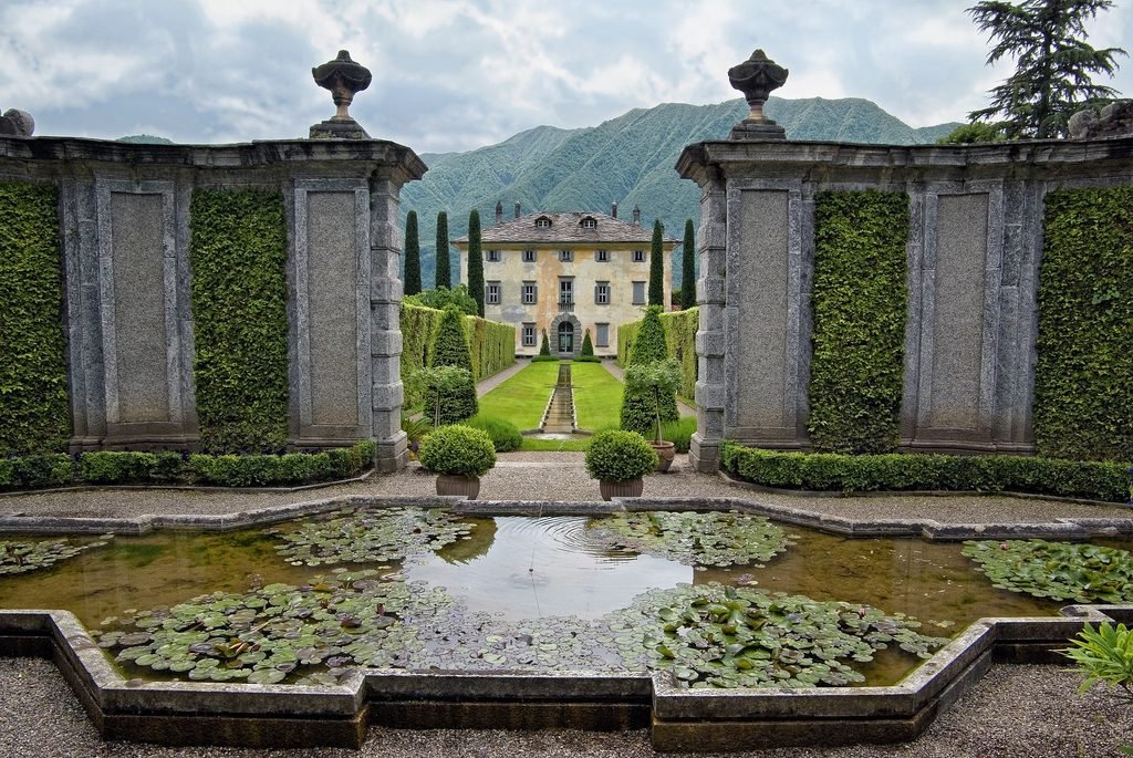 A symmetrical formal garden featuring a calm lily pond in the foreground, manicured green hedges, and stone walls leading to an elegant Italian-style villa with a mountain backdrop.