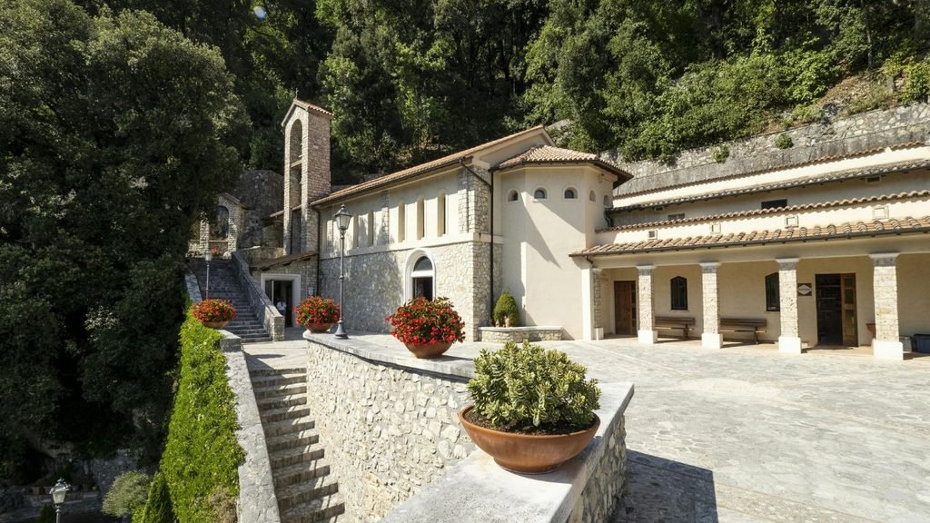 A historic stone church and bell tower with a terracotta tile roof, nestled into a lush, tree-covered hillside with stone stairs and potted red flowers in a sunny courtyard.