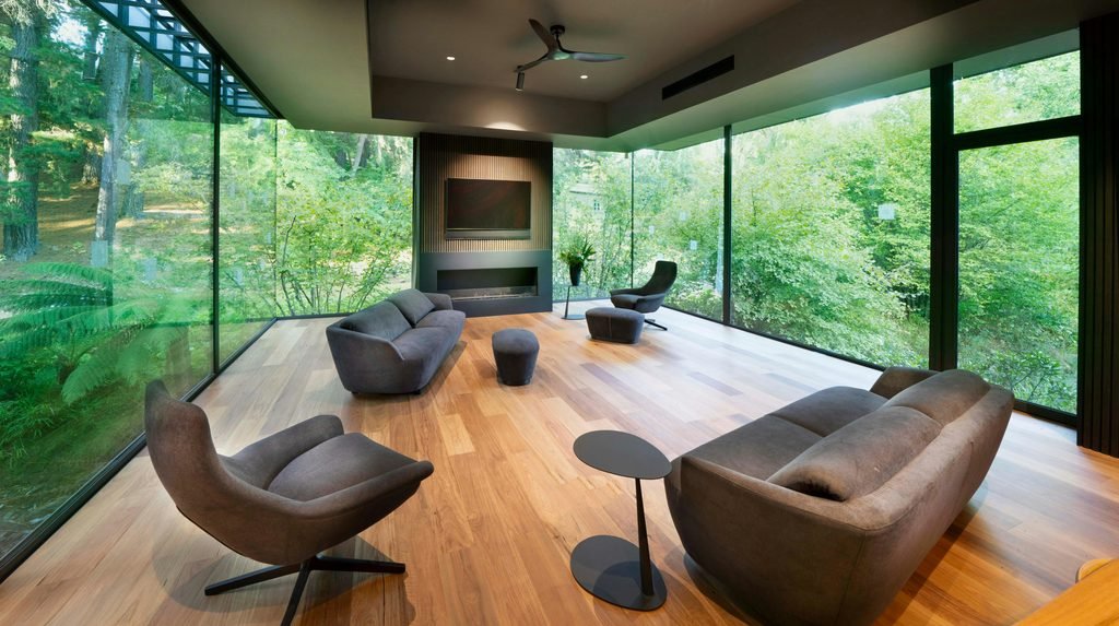 A luxury nature retreat featuring a minimalist modern living room with floor-to-ceiling glass walls, offering an immersive panoramic view of a lush green forest from sleek grey lounge seating.