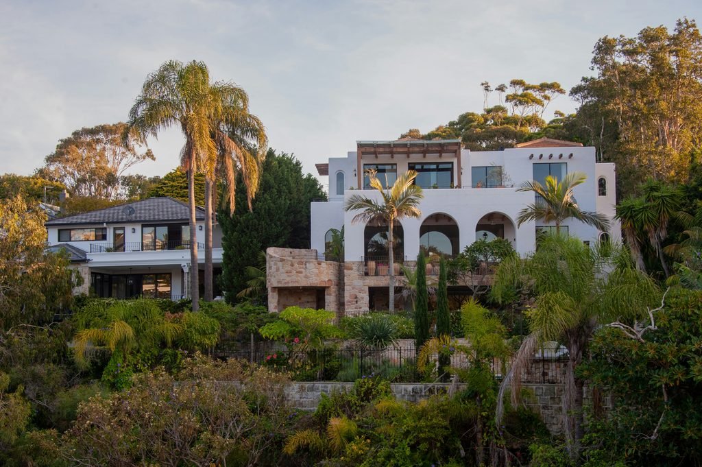 Luxurious multi-level white Mediterranean villa featuring elegant archways, expansive balconies, and stone retaining walls, surrounded by mature palm trees and lush tropical foliage.