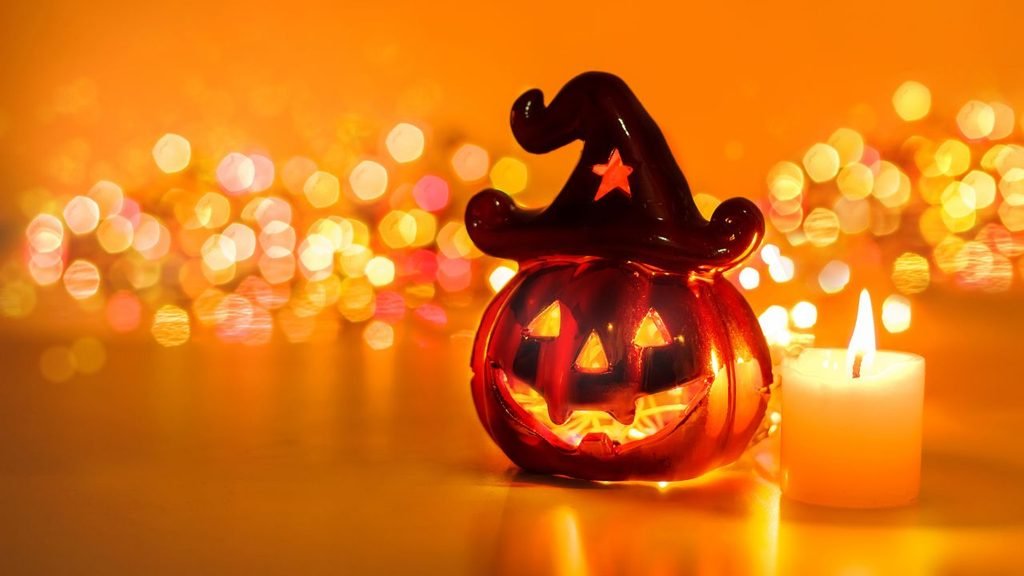 Small ceramic jack-o'-lantern decoration wearing a witch hat beside a lit candle with orange bokeh lights