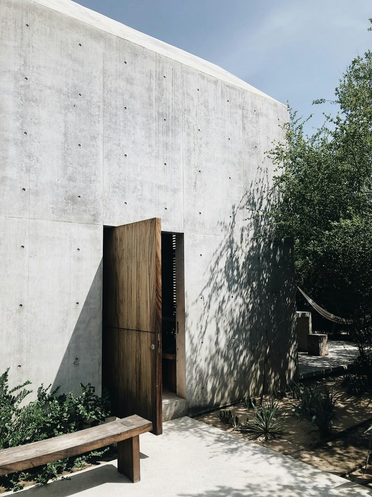 A modern, minimalist building made of smooth grey concrete featuring a massive, propped-open wooden door, a simple wooden bench, and drought-tolerant desert landscaping.