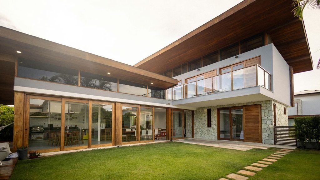 A modern, two-story home featuring a flat wooden overhanging roof, expansive glass sliding doors, stone accents, and a freshly manicured green lawn in the backyard.
