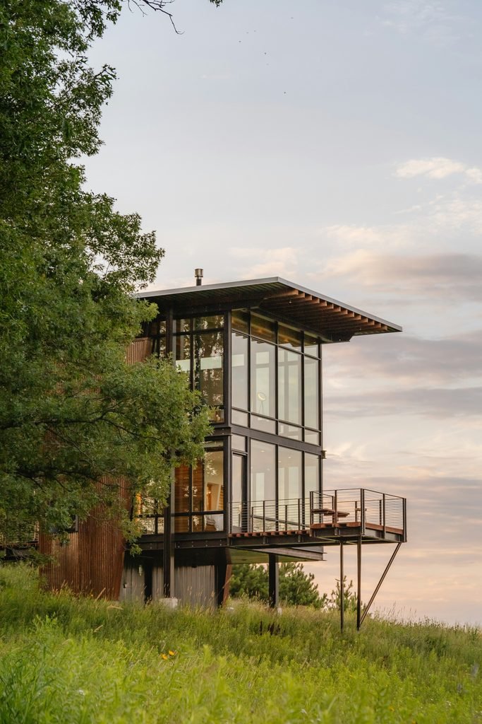 Modern multi-story glass and steel cabin perched on a grassy hillside, featuring an elevated observation deck and reflecting the warm colors of the sunset.