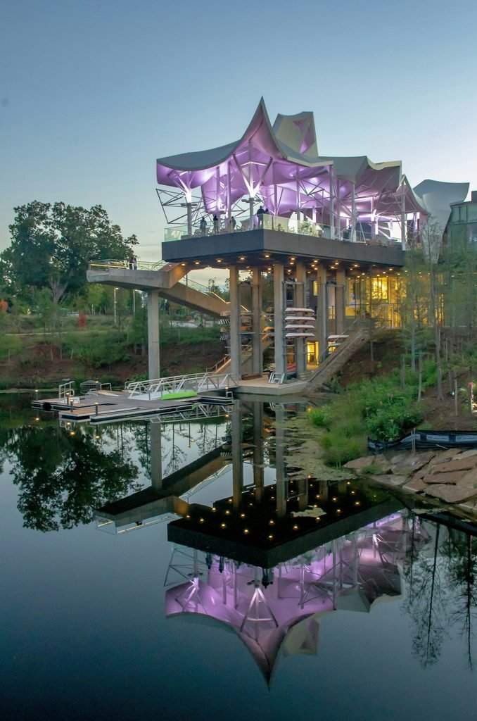 A modern waterfront pavilion and boathouse at dusk, featuring a striking illuminated purple canopy and tall concrete pillars, with its reflection mirrored perfectly in the calm water above a floating dock.