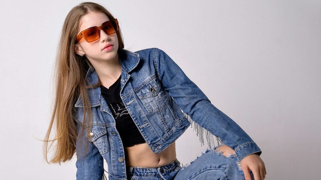 Model wearing an orange-tinted sunglasses and a cropped denim jacket in a studio portrait