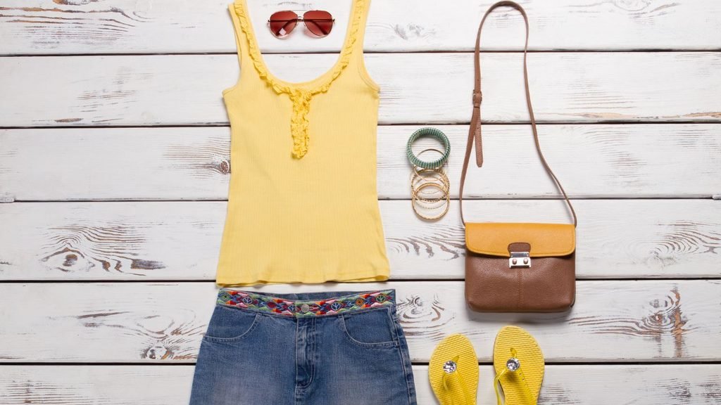 A flat lay of a women's summer outfit on a white wooden background, featuring a yellow tank top, denim skirt with an embroidered belt, sunglasses, bracelets, a brown crossbody bag, and yellow flip-flops.