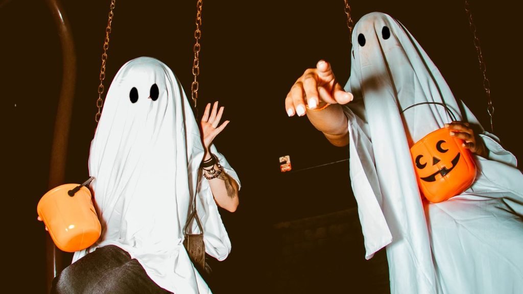 Two people in white ghost costumes swinging on a playground at night holding orange candy buckets
