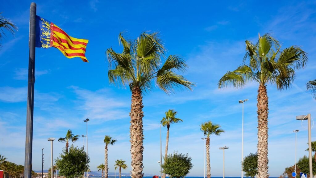 Flag of the Valencian Community waving above palm trees near the beach promenade