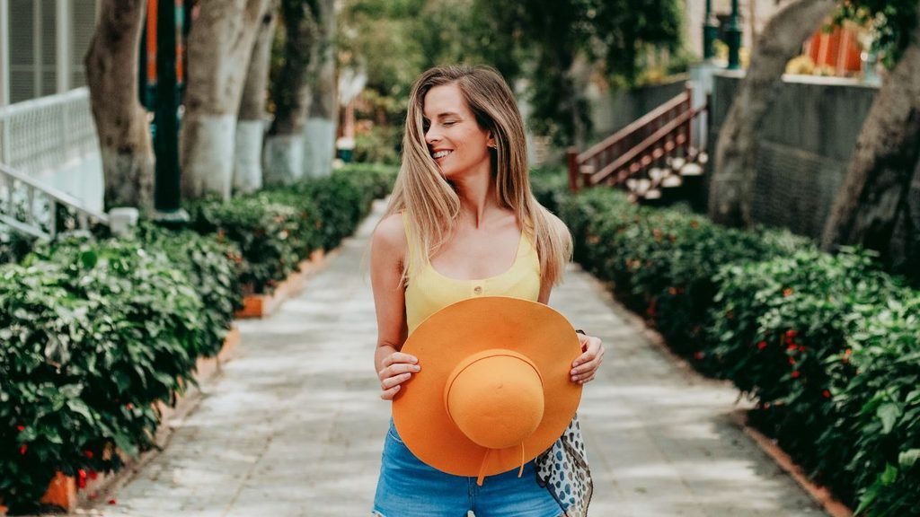 A smiling woman holding an orange wide-brimmed hat, wearing a yellow tank top and denim shorts, walking along a sunny, tree-lined path.