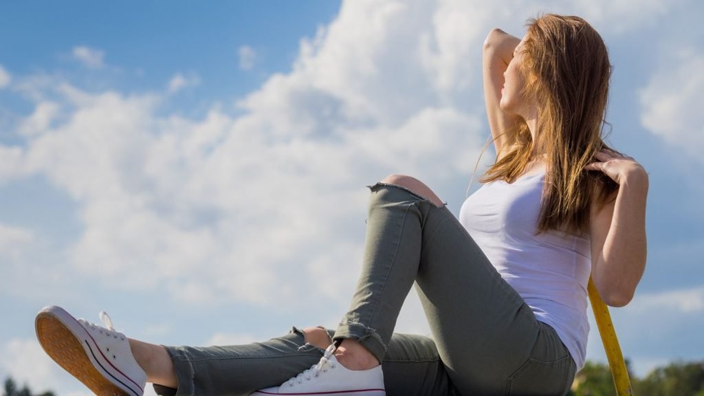 Woman in a white tank top and ripped jeans sitting relaxed against a blue sky backdrop