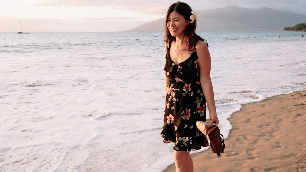 A happy woman wearing a black floral dress and a white flower in her hair walking barefoot on a sandy beach at sunset, carrying her sandals.
