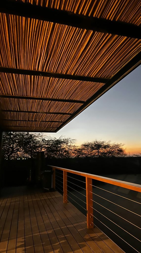 Tranquil outdoor wooden deck featuring a rustic bamboo canopy ceiling, looking out over silhouetted trees against a vibrant sunset sky.
